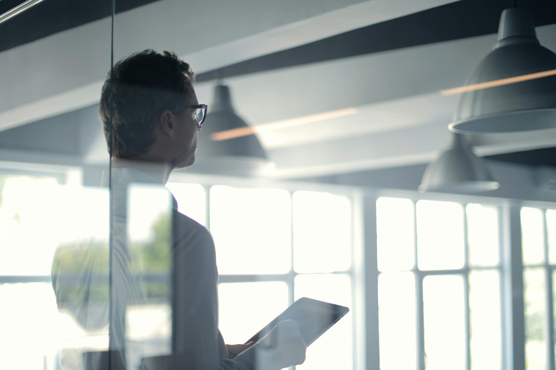 A businessman holding a tablet in an office, looking thoughtfully through a glass wall.